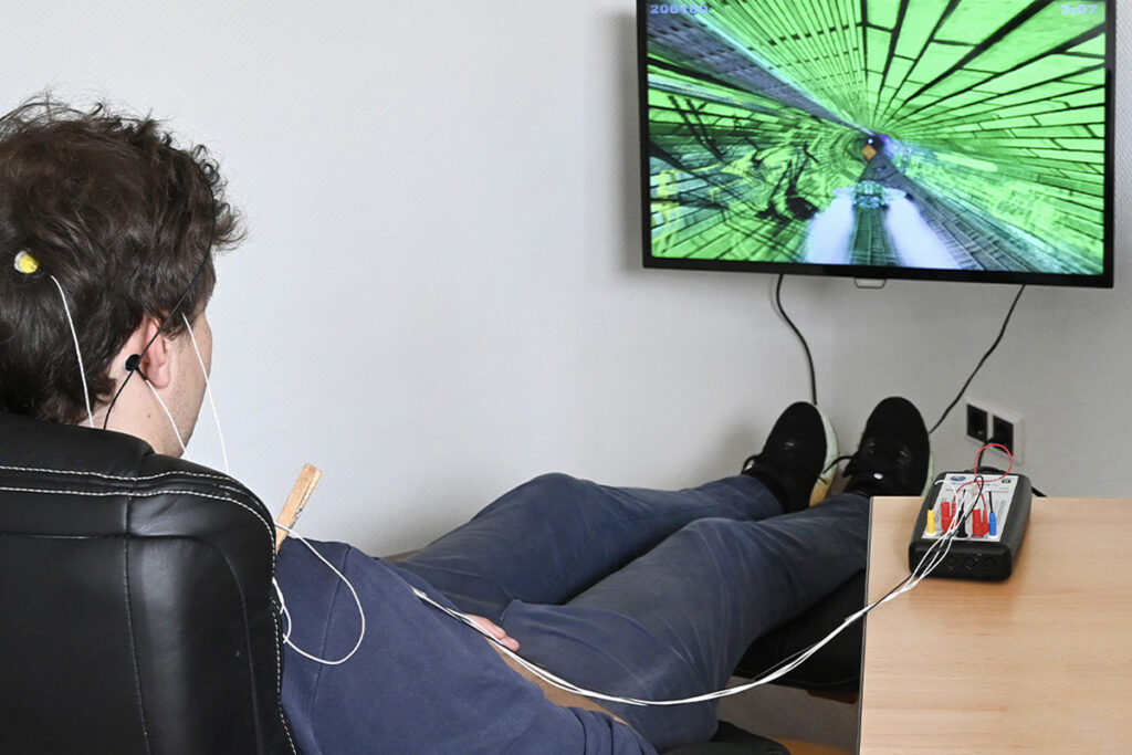Junger Mann mit Elektroden am Kopf schaut auf einen Fernseher. Dieser zeigt einen Flugsimulator in grünem Tunnel