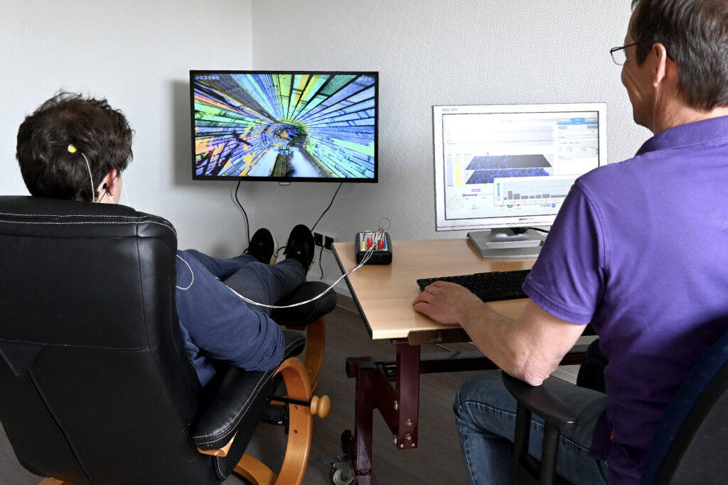 Mann an Computer und junger Mann mit Elektroden vor einem Fernseher.