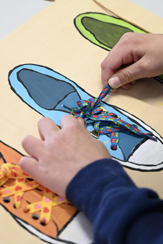 Auf Holzbrett gemalter Schuh. Kinderhände machen darauf eine Schleife in die Schnürsenkel.